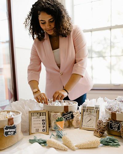 A woman in a pink blazer stands behind a table displayed with baked goods.